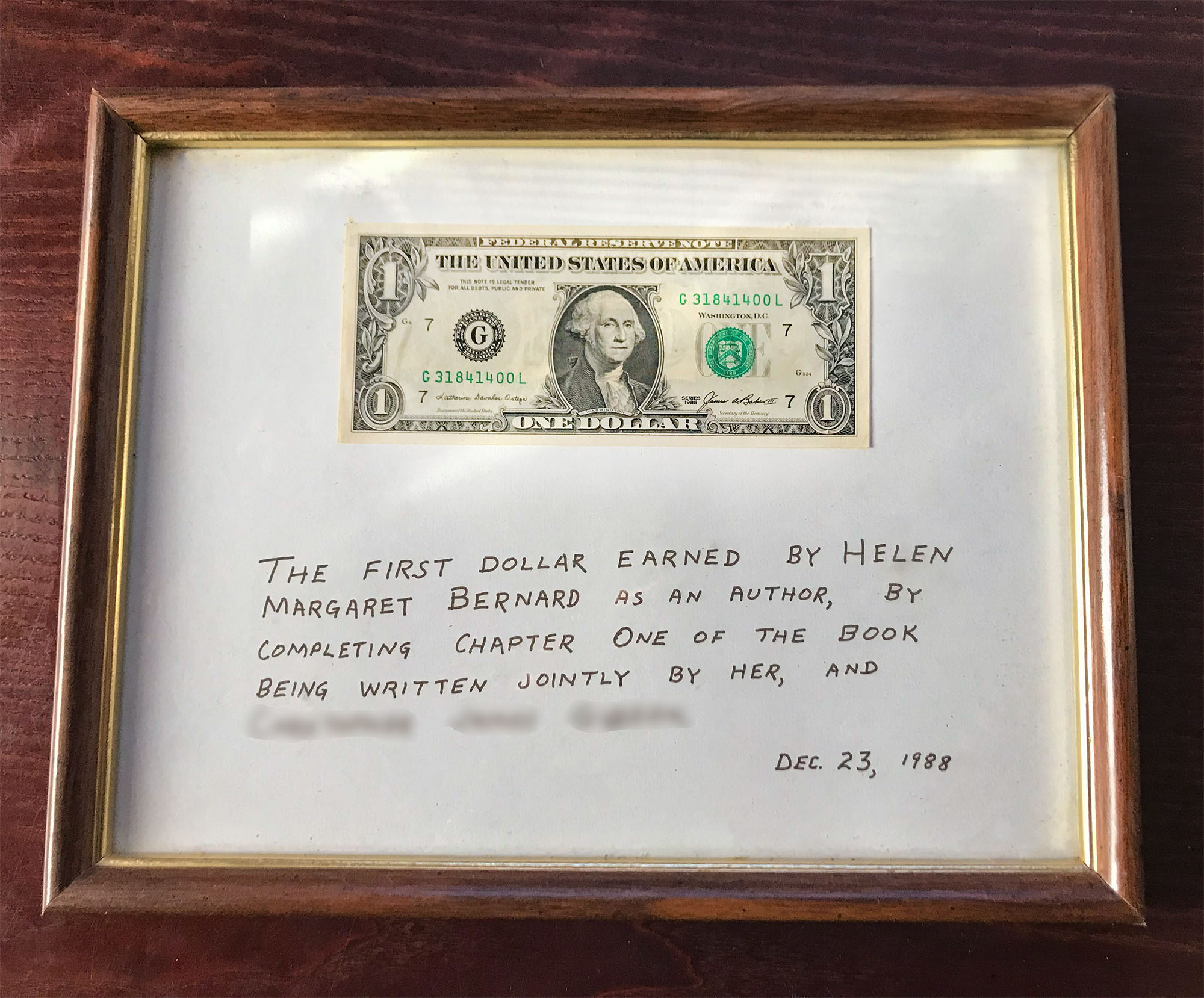 Framed dollar bill with the words, 'The first dollar earned by Helen Margaret Bernard as an author, by completing chapter one of the book being written jointly by her, and (name blurred for privacy reasons) December 23rd, 1988.